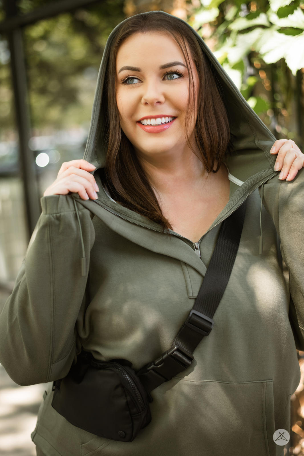 Smiling woman outdoors wearing olive SweetTops All Day Hoodie with hood up and crossbody bag in natural light