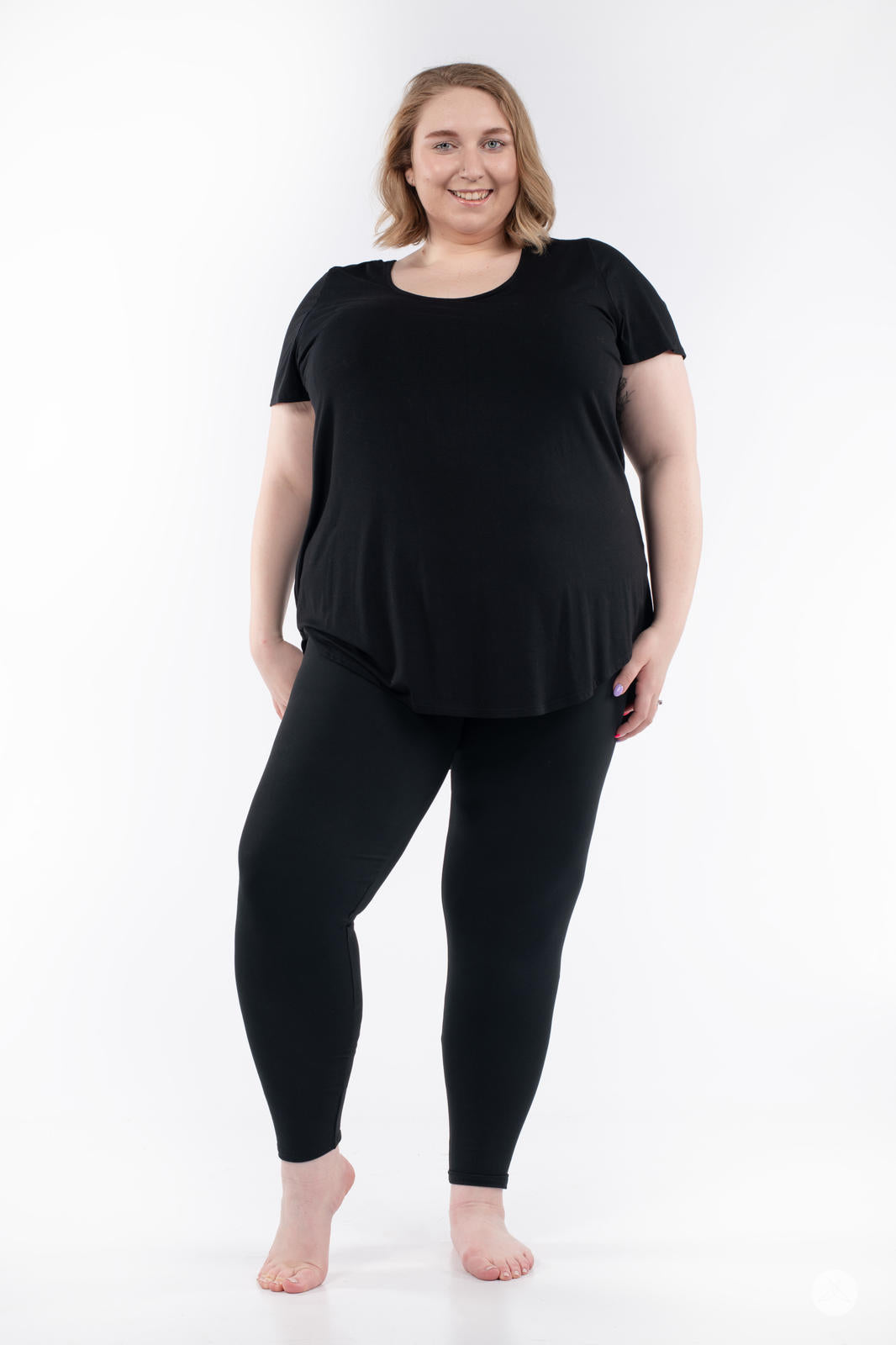 Smiling plus-size model wearing SweetLegs Black Licorice leggings and matching black top barefoot on white background
