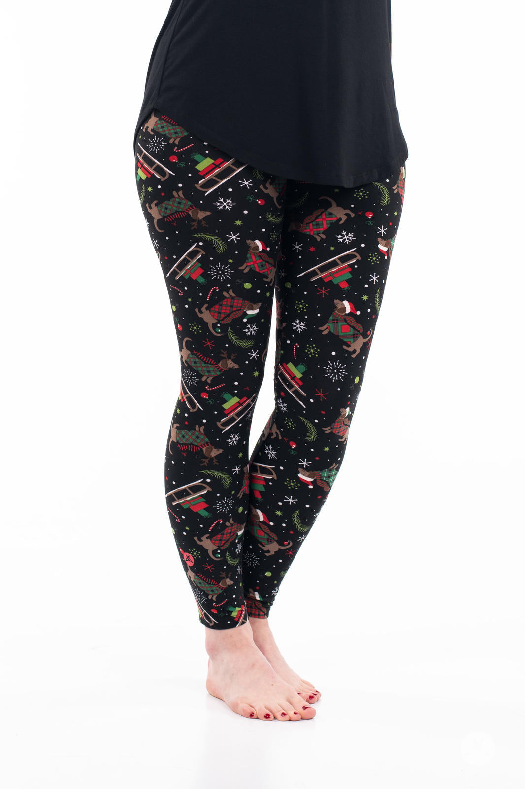 Adult wearing SweetLegs Happy Pawlidays leggings with dachshunds in holiday attire, sleighs, and snowflakes on black background