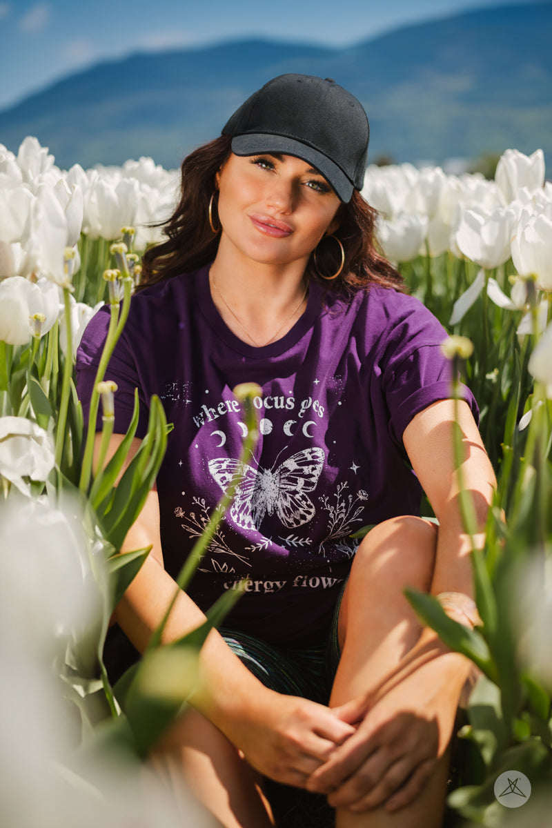 Woman sitting among white tulips wearing SweetTops Vintage Graphic Tee in purple with mystical butterfly design and affirmations
