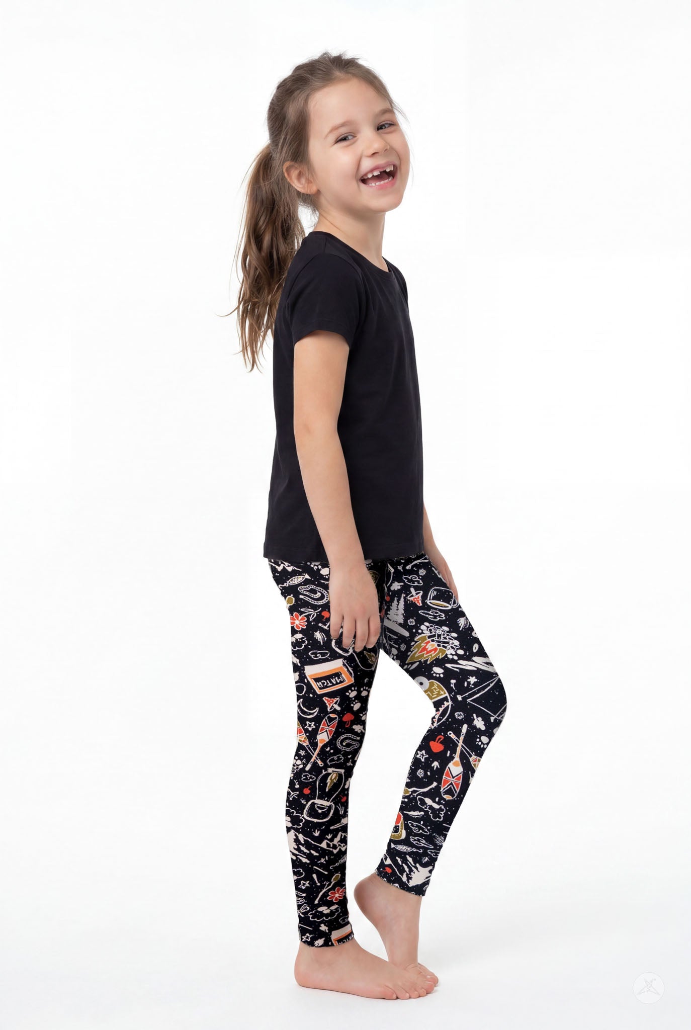 Child wearing SweetLegs Off Grid Kids patterned leggings and black tee standing barefoot smiling side view