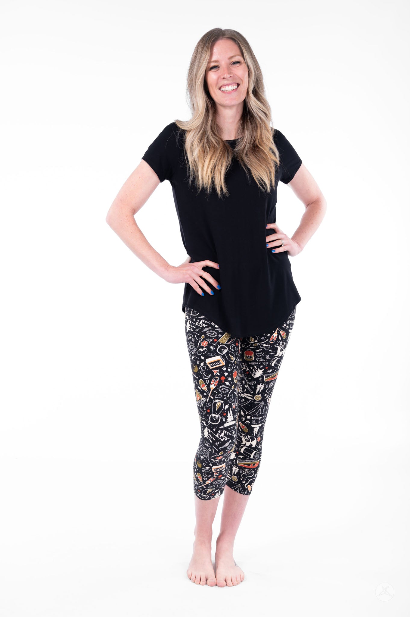 Woman smiling wearing SweetLegs Off Grid Crops with colorful outdoor-themed pattern and black top standing barefoot front view