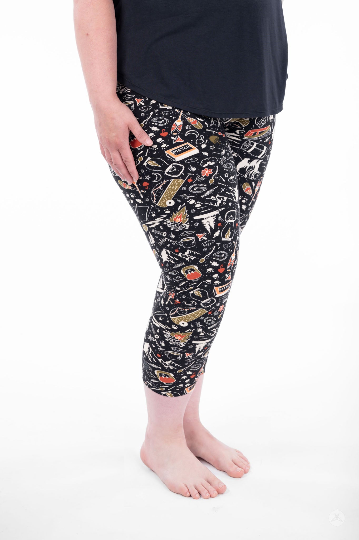 Plus size model wearing SweetLegs Off Grid Crops with camping pattern in black standing barefoot side view
