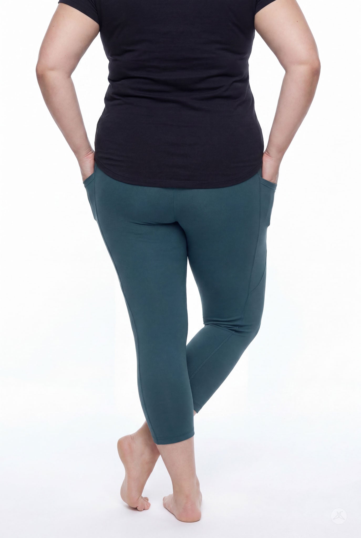 Plus size model wearing SweetLegs Deep Teal Pocket Crops leggings standing barefoot with hands in side pockets back view