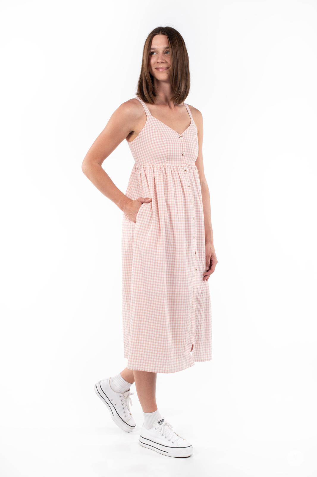 Model wearing SweetTops Gingham Dress in soft pink with button front, pockets, and adjustable straps standing on white background