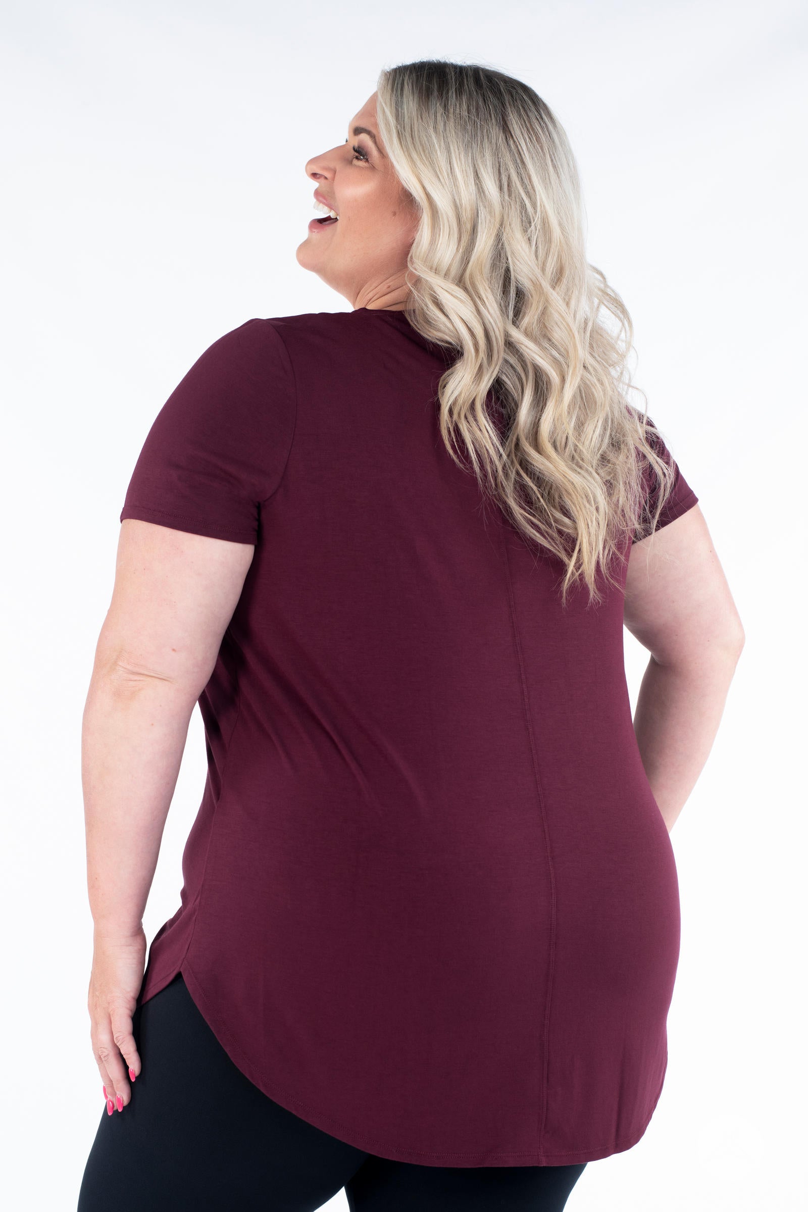 Plus size woman smiling wearing maroon Classic Tee by SweetLegs Canada with short sleeves and high-low hem back view