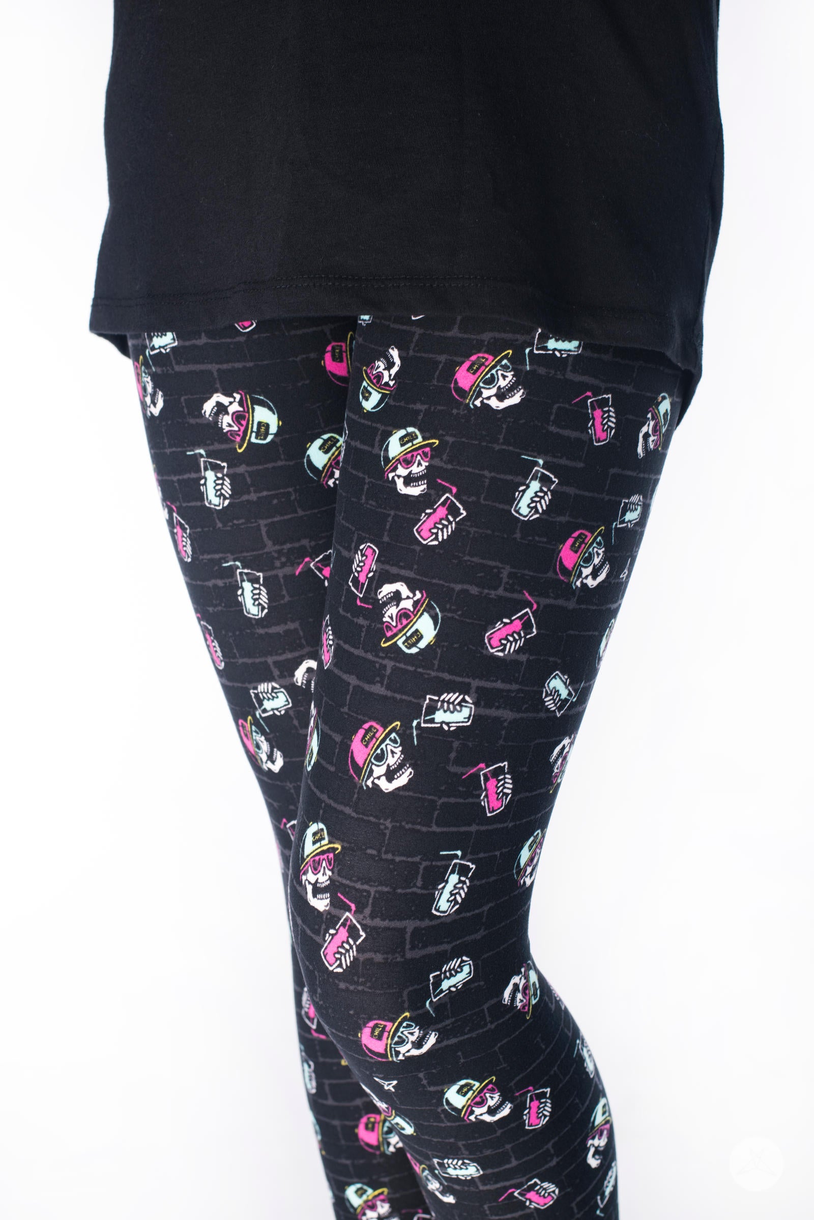 SweetLegs Stay Chill Kids leggings with pink and mint cartoon skulls wearing hats and sipping drinks on black brick background