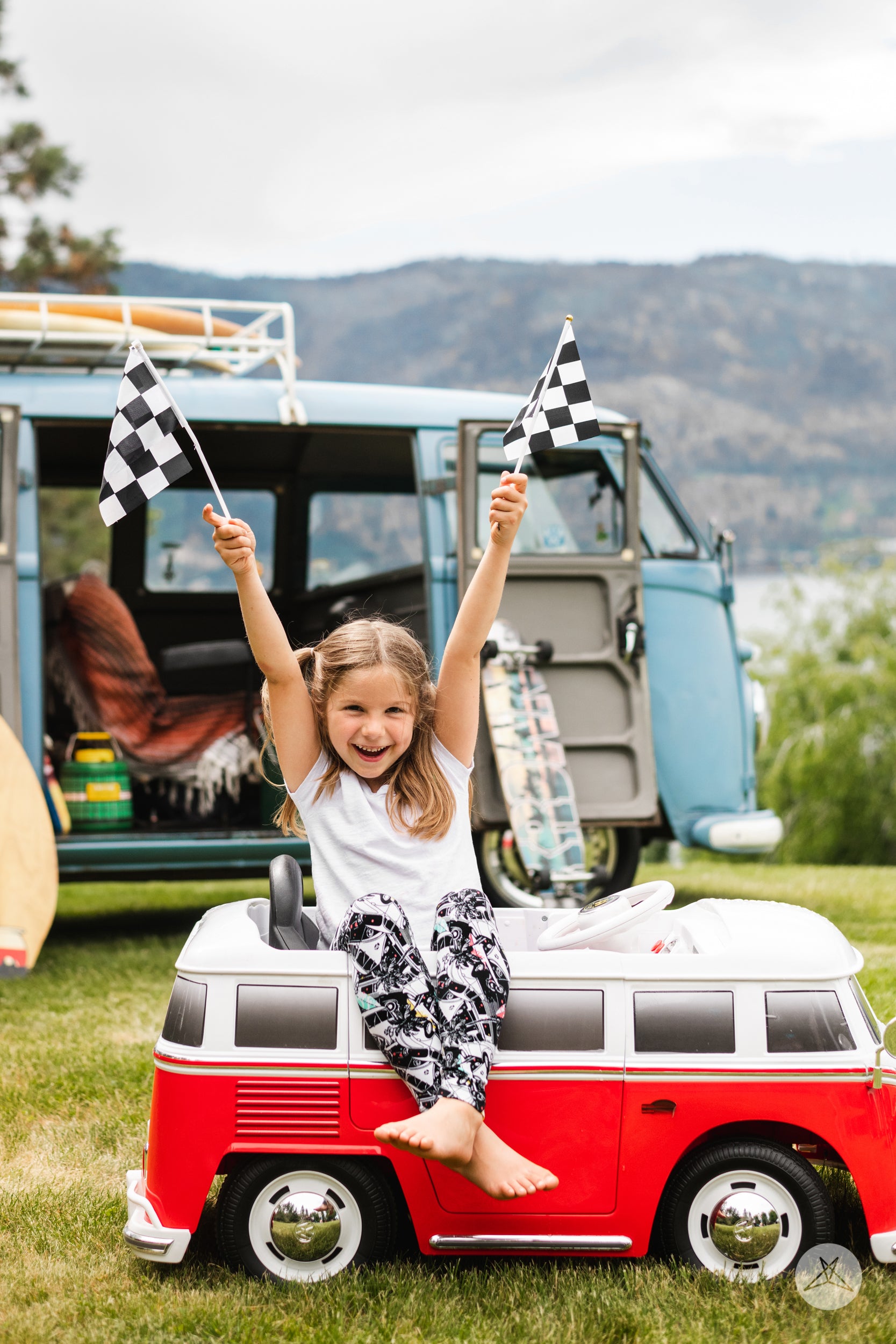 Girl wearing California Dreamin' Kids by SweetLegs sitting by red toy van holding two checkered flags outdoors
