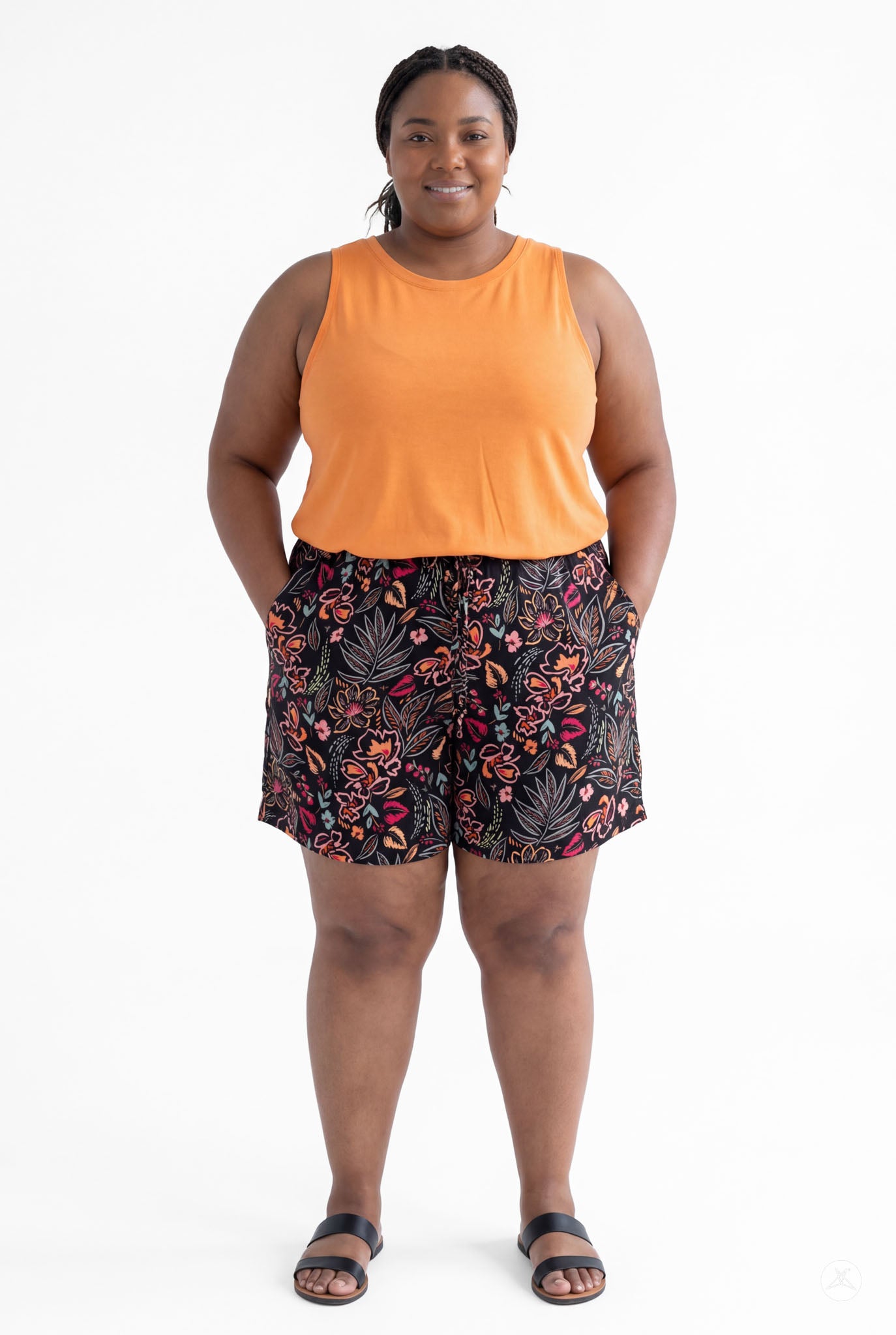 Plus size model wearing SweetLegs Breeze Shorts with floral print and orange sleeveless top, hands in pockets, smiling