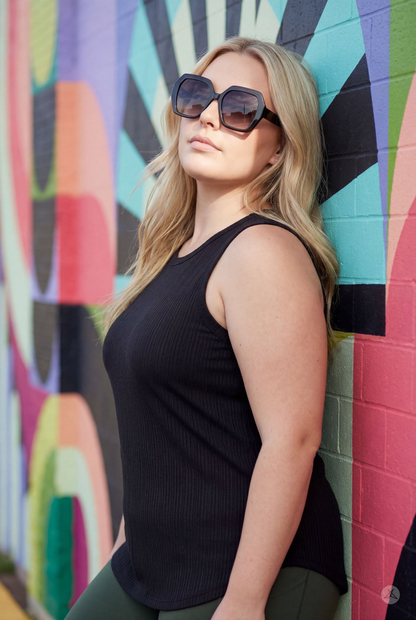 Woman wearing The Soft Ribbed Tank by SweetTops in black, leaning against colorful mural with oversized sunglasses