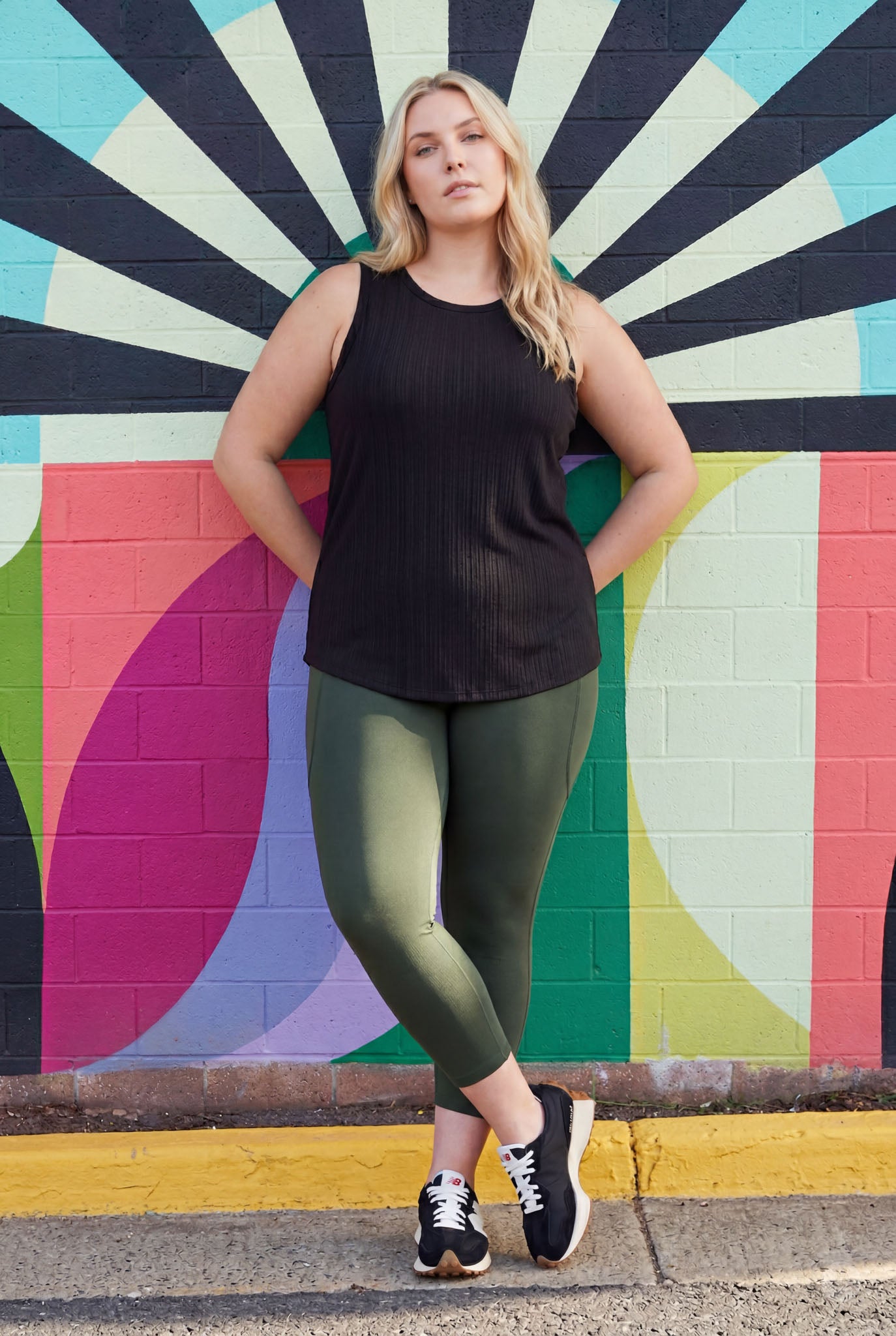 Woman standing in front of colorful mural wearing SweetLegs Cedar Pocket Crops in olive green with black sleeveless top