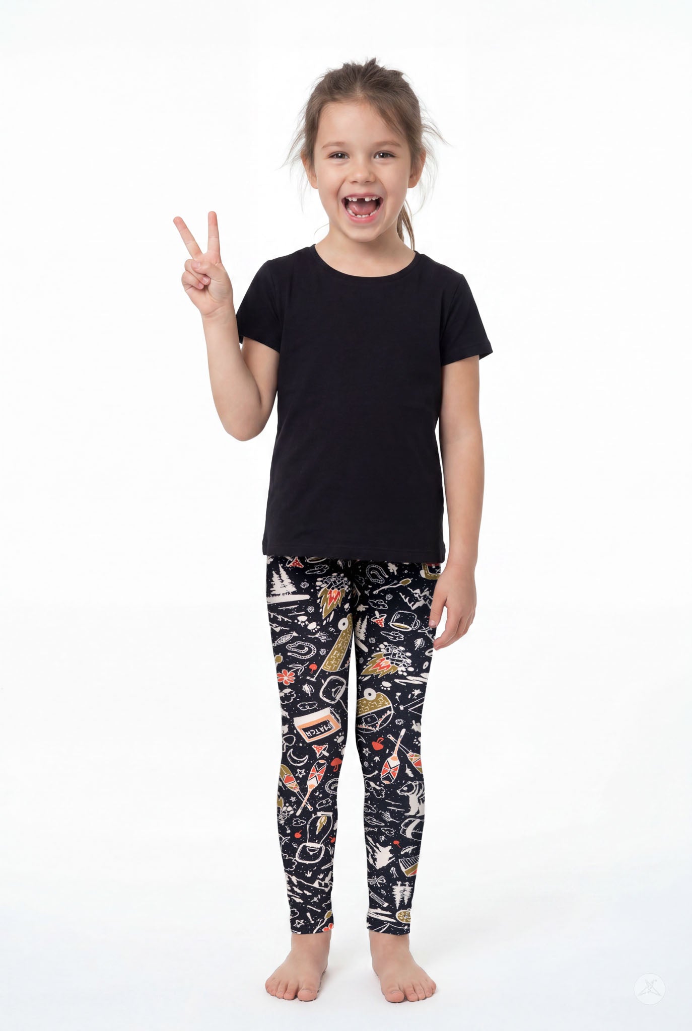 Smiling child wearing SweetLegs Off Grid Kids patterned leggings and black tee standing barefoot showing peace sign front view