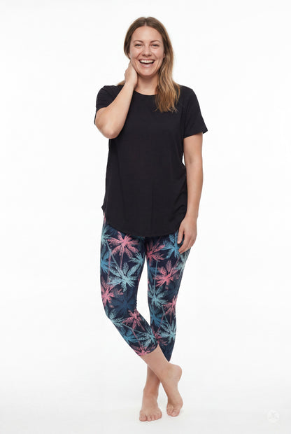 Smiling woman standing barefoot wearing SweetLegs Tiki Nights Crops with blue and pink palm tree pattern and black tee