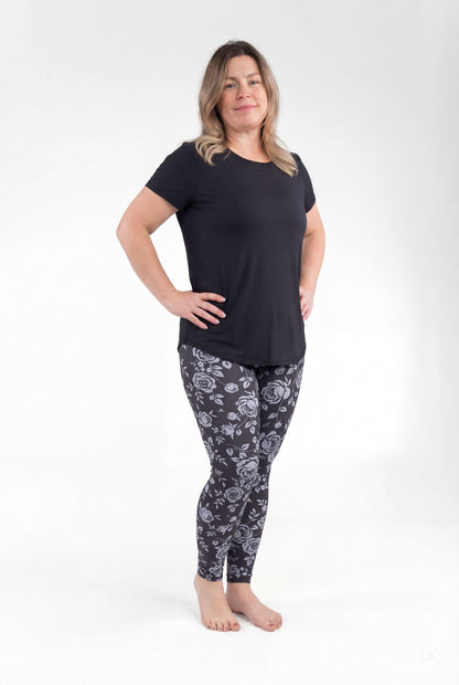 Woman wearing SweetLegs Shadow Garden leggings with grey-blue floral print on dark background, standing barefoot confidently