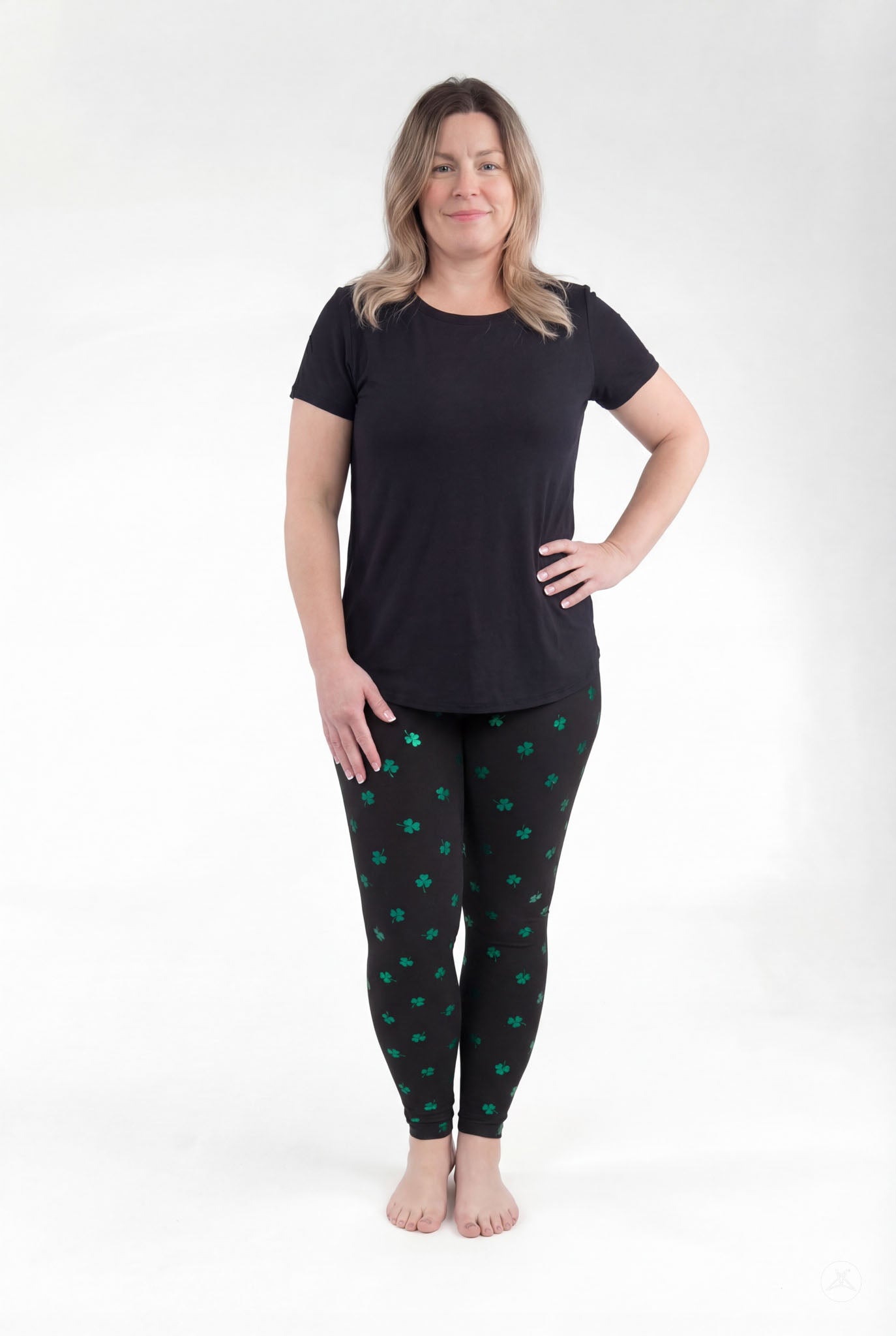 Woman modeling SweetLegs Lucky Glow leggings with green shamrock print on black background standing barefoot