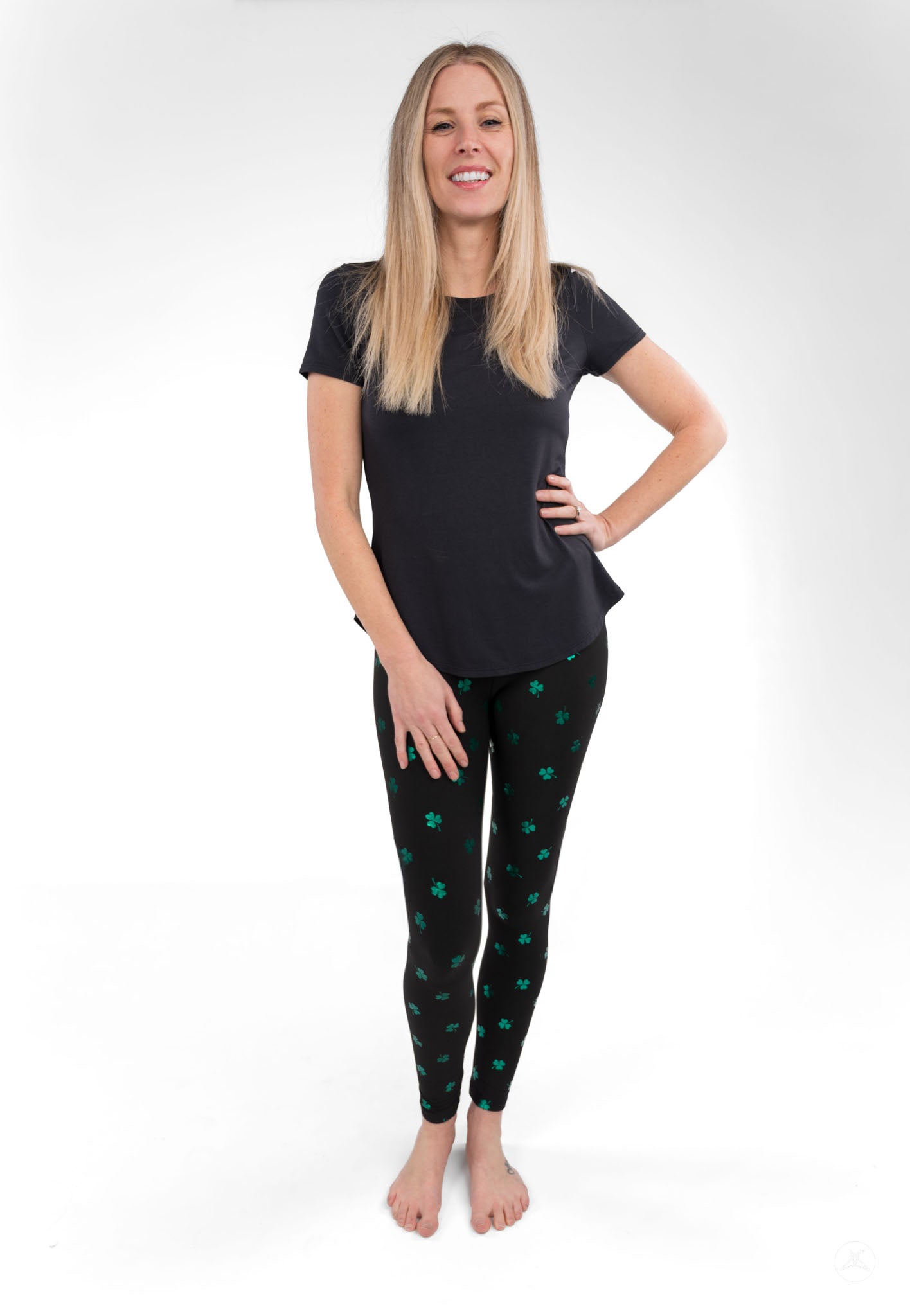 Woman wearing SweetLegs Lucky Glow leggings with emerald shamrock foil accents on a dark background smiling confidently