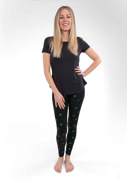Woman wearing SweetLegs Lucky Glow leggings with emerald shamrock foil accents on a dark background smiling confidently